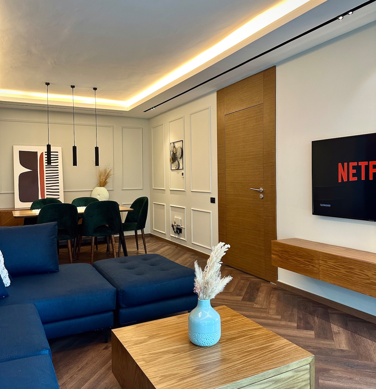 A modern living area features a dark sectional sofa and a wooden coffee table. A flat-screen TV mounted on the wall displays the Netflix logo. Adjacent is a dining area with a table surrounded by green chairs, accented by wall art and soft overhead lighting.