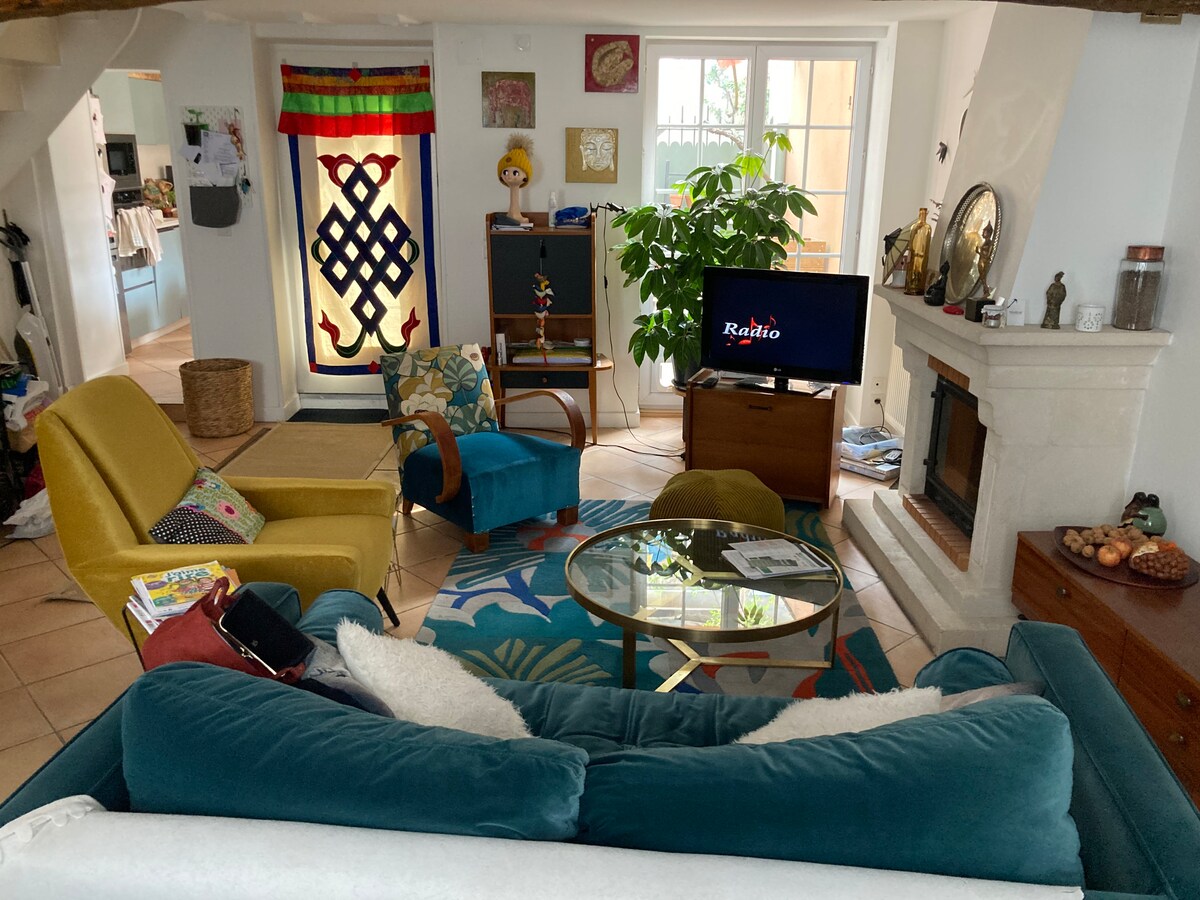 A cozy living area is showcased with a plush teal sofa and a glass coffee table at its center. Colorful décor elements, including a vibrant rug and wall hangings, are present. A decorative fireplace adds character, while a large plant enhances the space. A television is positioned nearby.