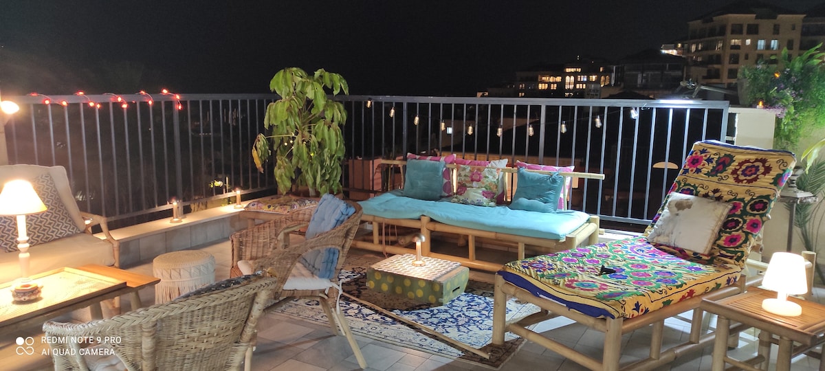A rooftop terrace is showcased at night, featuring a mix of seating options including a daybed and lounge chairs with vibrant cushions. Soft lighting and decorative plants create a cozy atmosphere, while a view of the surrounding buildings is visible in the background.