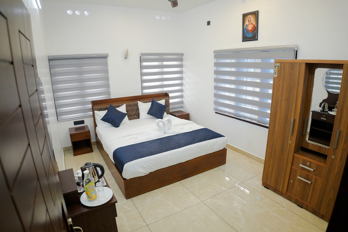 A well-appointed bedroom is showcased with a spacious bed featuring decorative pillows. Natural light enters through two window shades. A stylish wooden wardrobe and a nightstand with amenities are included, enhancing the room's functionality.