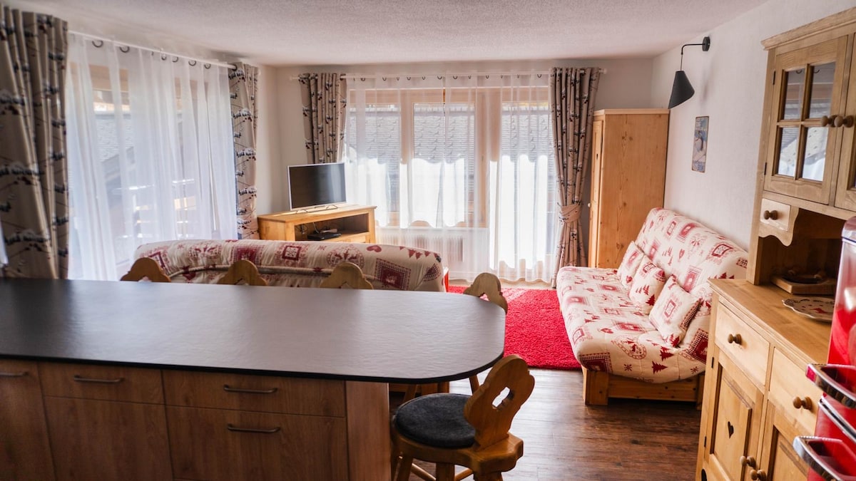 A cozy living area is arranged with two patterned sofas and a wooden coffee table. Natural light filters through sheer curtains, illuminating the space. A well-equipped kitchen features wooden cabinetry and a bar area, complemented by a vibrant red rug that adds a touch of warmth.