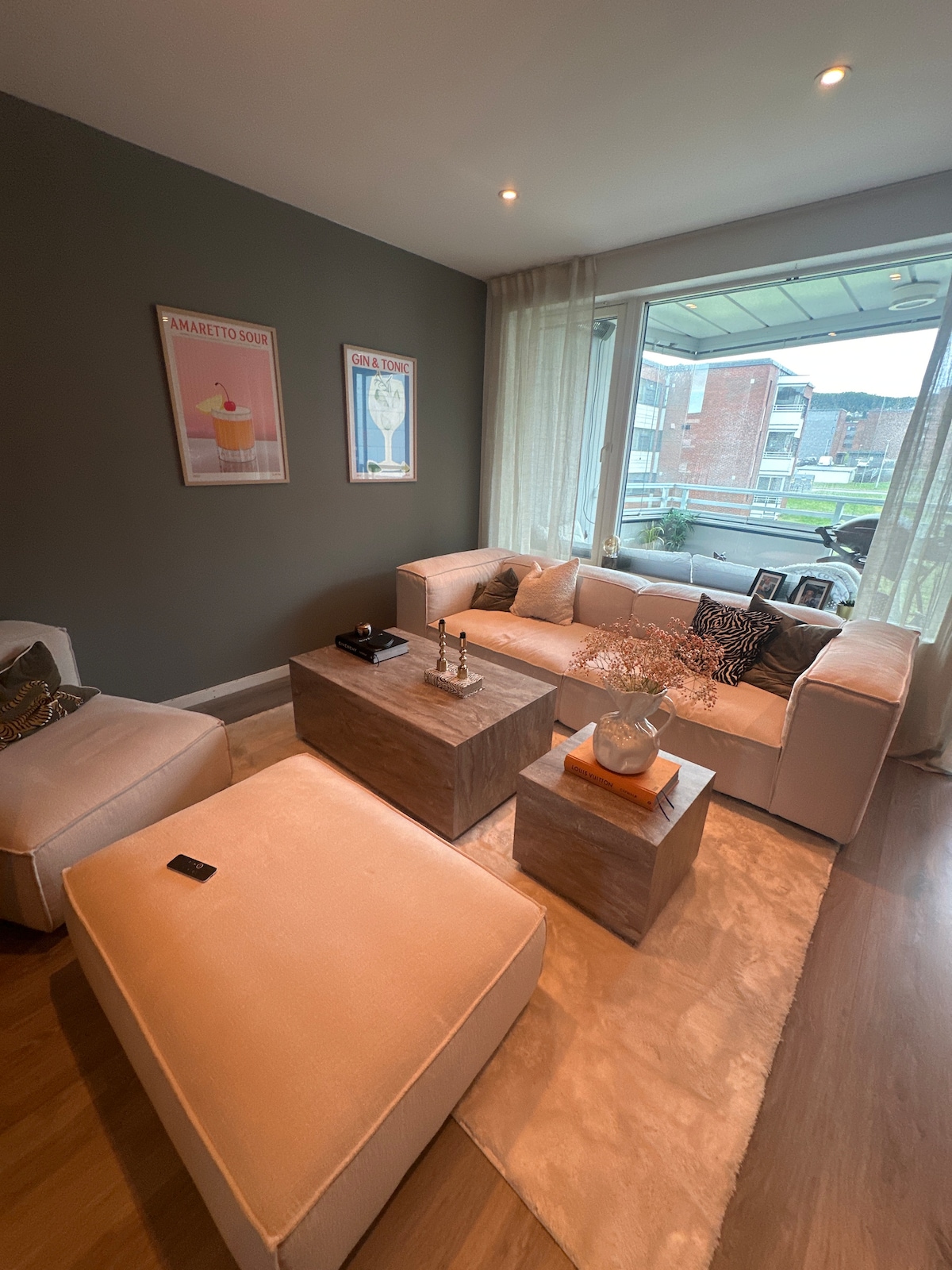 A modern living area features a light-colored sectional sofa arranged around a wooden coffee table. Two framed art pieces adorn the wall, while a plush area rug softens the floor. A large window allows natural light to fill the space, with greenery visible outside.