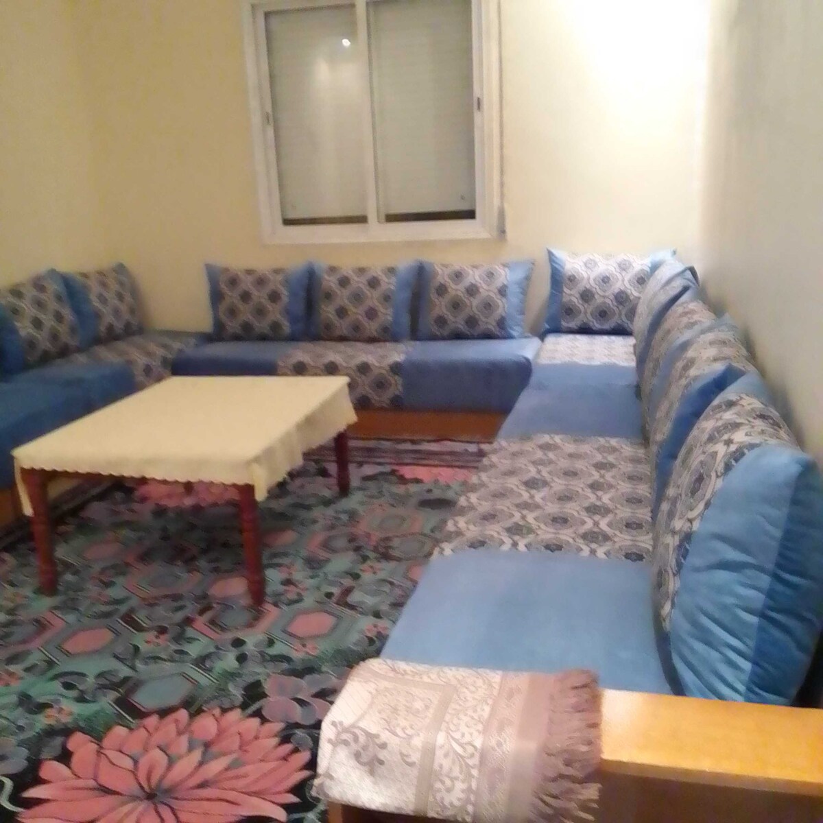 A comfortable seating area features a spacious L-shaped couch with blue and patterned cushions. A low wooden table is centered on a vibrantly patterned rug, while light enters through a window adorned with light-colored blinds.