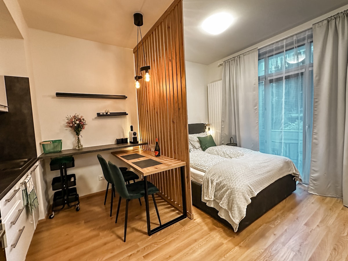 A well-equipped living space features a spacious bed with light bedding and decorative cushions. A wooden partition separates the bed area from a small dining table with four chairs. Natural light enters through large curtains, enhancing the calm atmosphere.