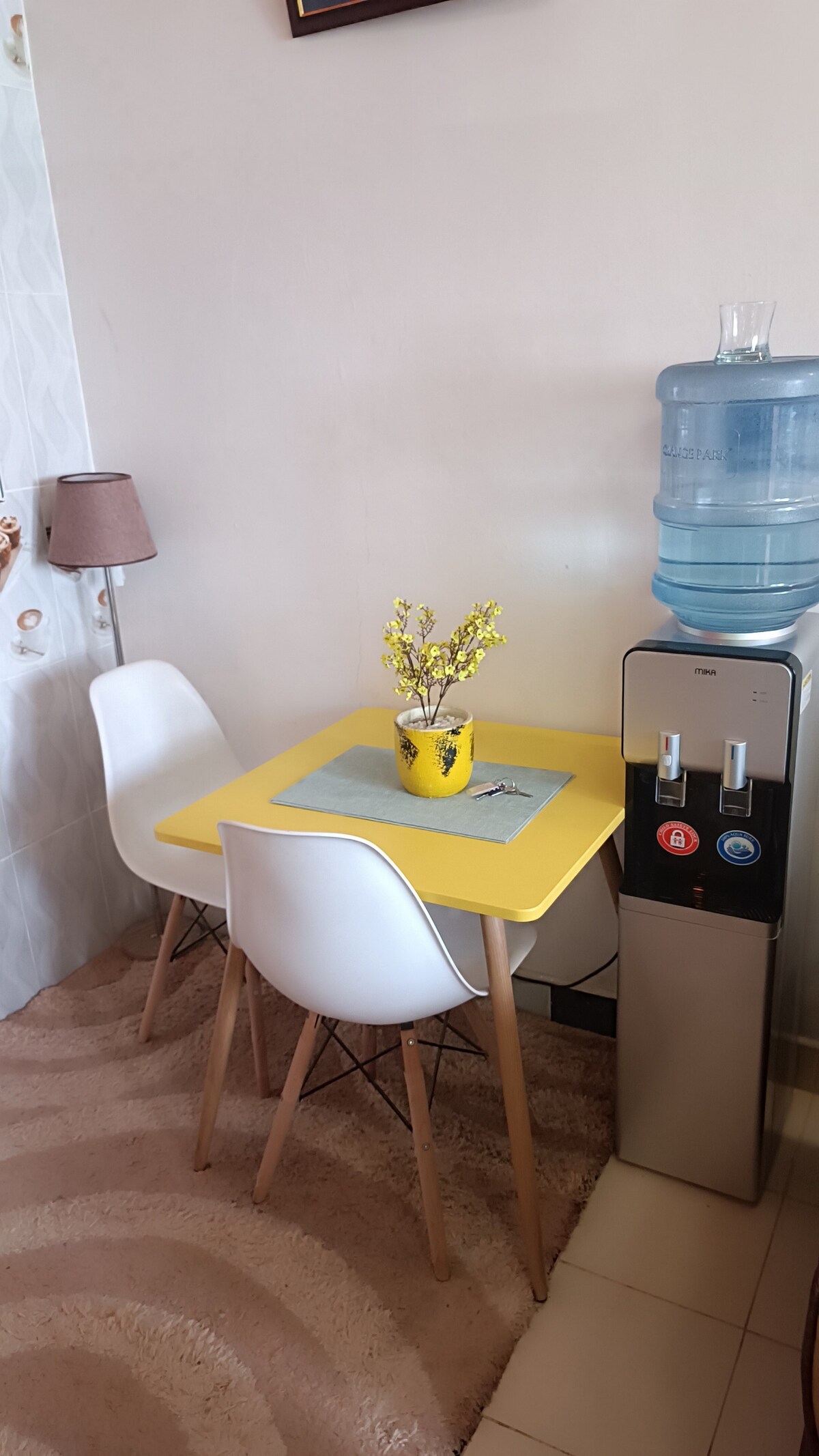 A small dining area features a square yellow table complemented by two white chairs. A decorative yellow pot with flowers sits atop the table, while a water cooler stands nearby, providing convenience. Soft lighting from a lamp on the wall creates a cozy ambiance.
