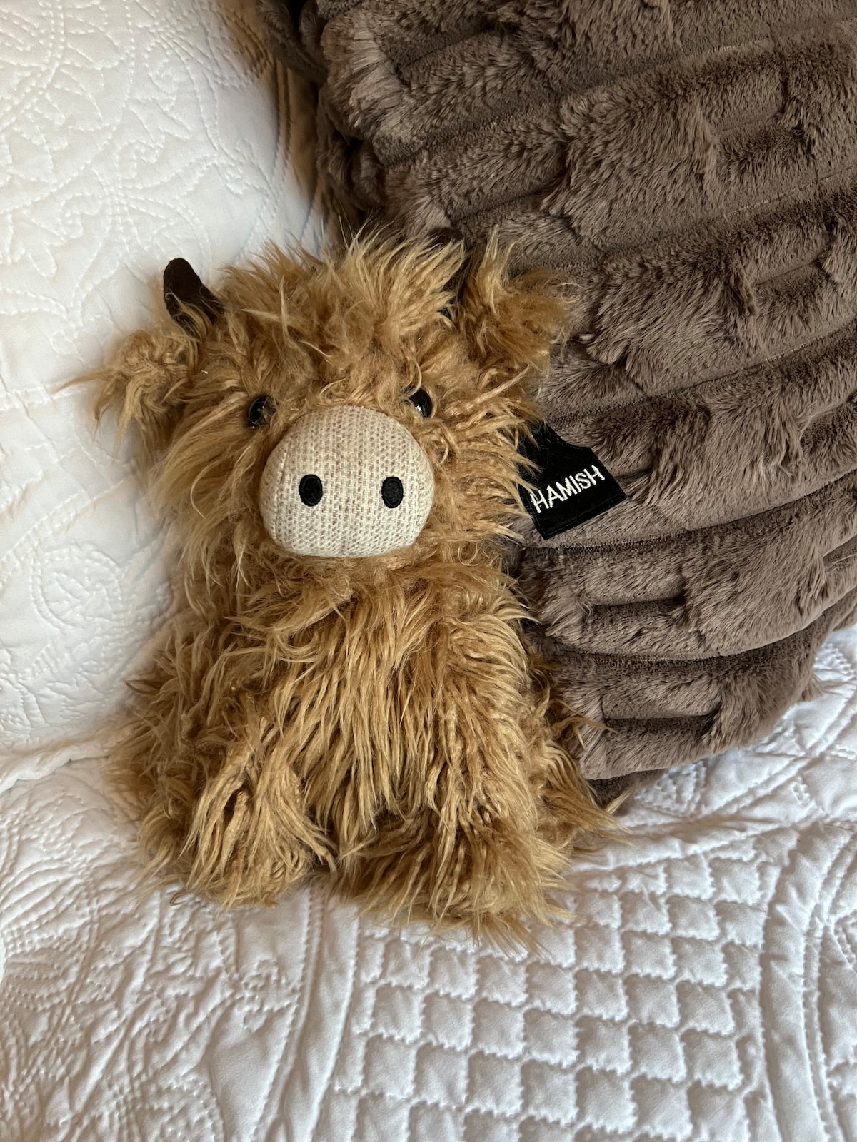 A soft plush toy resembling a highland cow is positioned on a white quilted bedspread, accompanied by a cozy brown throw pillow. The toy features a fuzzy texture with small horns and a textured nose, adding a playful element to the serene bedroom setting.