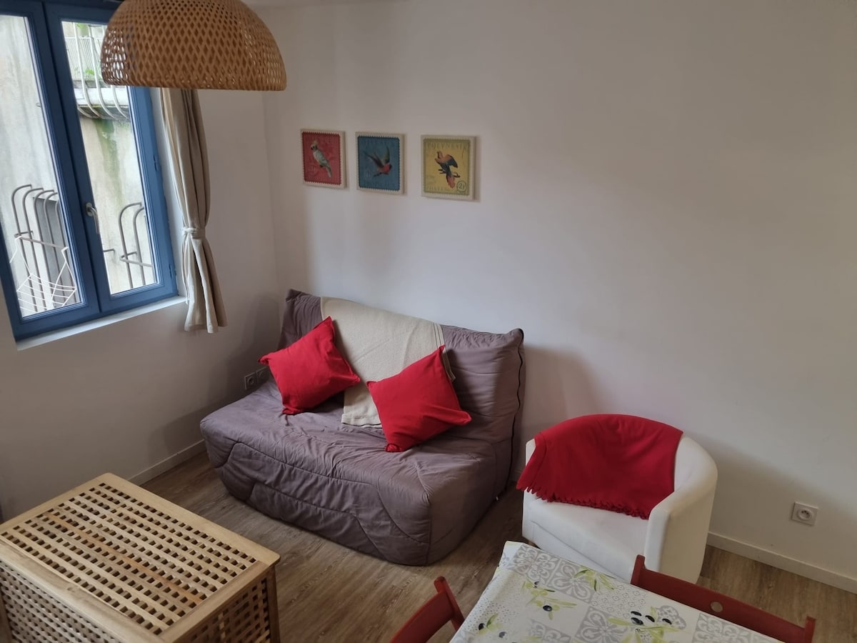 A comfortable living area includes a convertible sofa adorned with red cushions and a white armchair. A wooden coffee table with a woven design is positioned in front. Four framed artworks hang on the wall, and natural light enters through a window with light drapes.