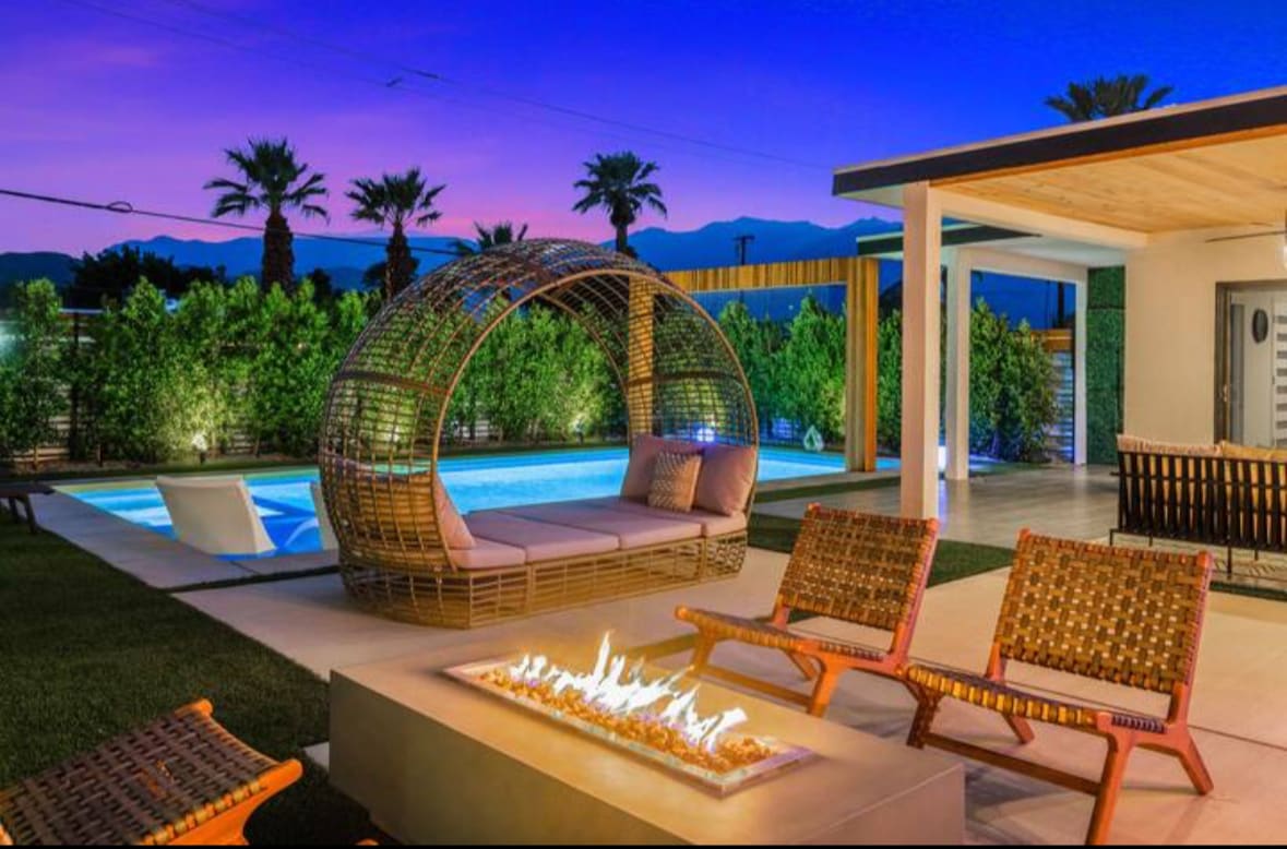A tranquil outdoor space features a heated pool under a vibrant sunset sky. The comfortable lounge area includes a circular seating structure and two wooden chairs surrounding a fire pit, all complemented by lush hedges and palm trees in the background.