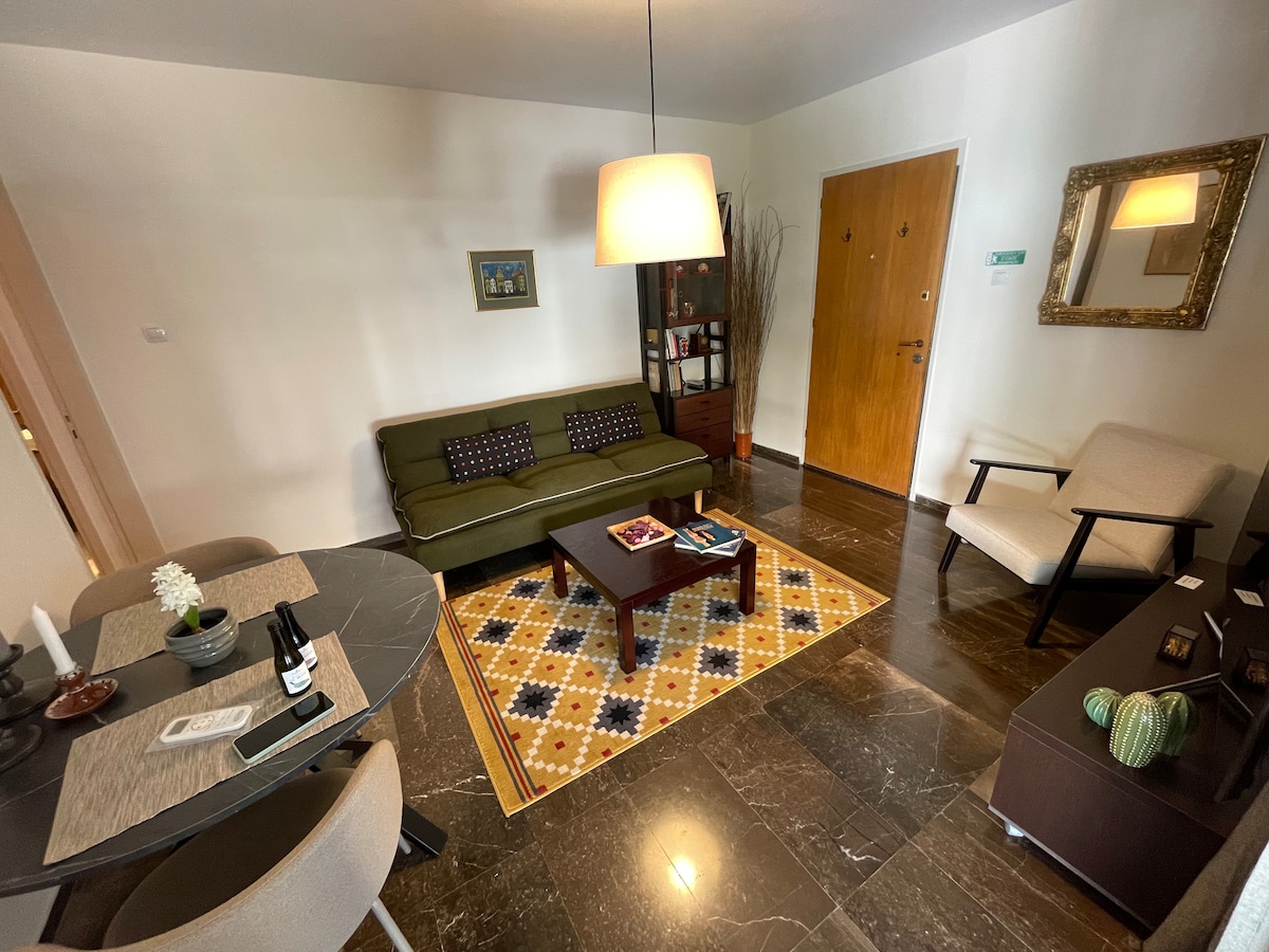 A living area features a green sofa with decorative cushions, a dark wooden coffee table, and a patterned area rug. Natural light enters through a nearby window. A small dining table with two chairs is positioned to the left, and a decorative mirror adds dimension to the space.