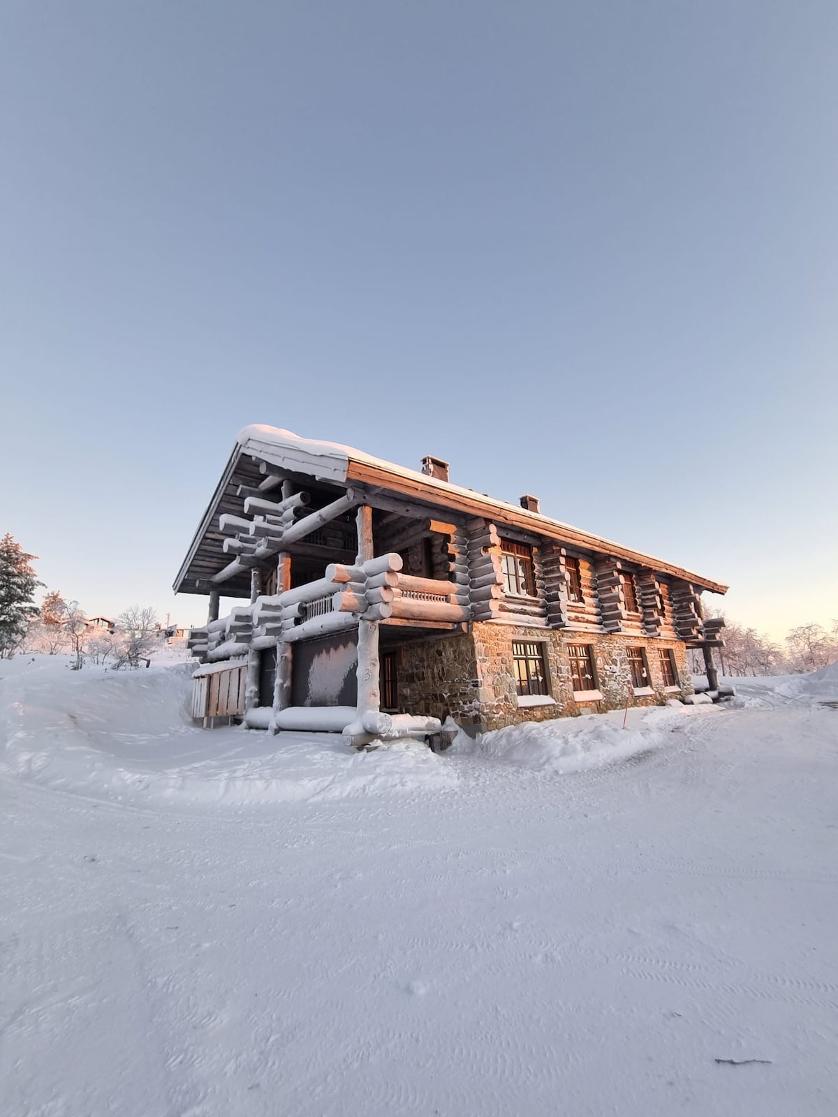 A charming log cabin is depicted, set against a serene winter backdrop. Snow covers the roof and ground, while frosted trees surround the structure. Large windows allow natural light to fill the interior, and the rustic design integrates well with the surrounding landscape.