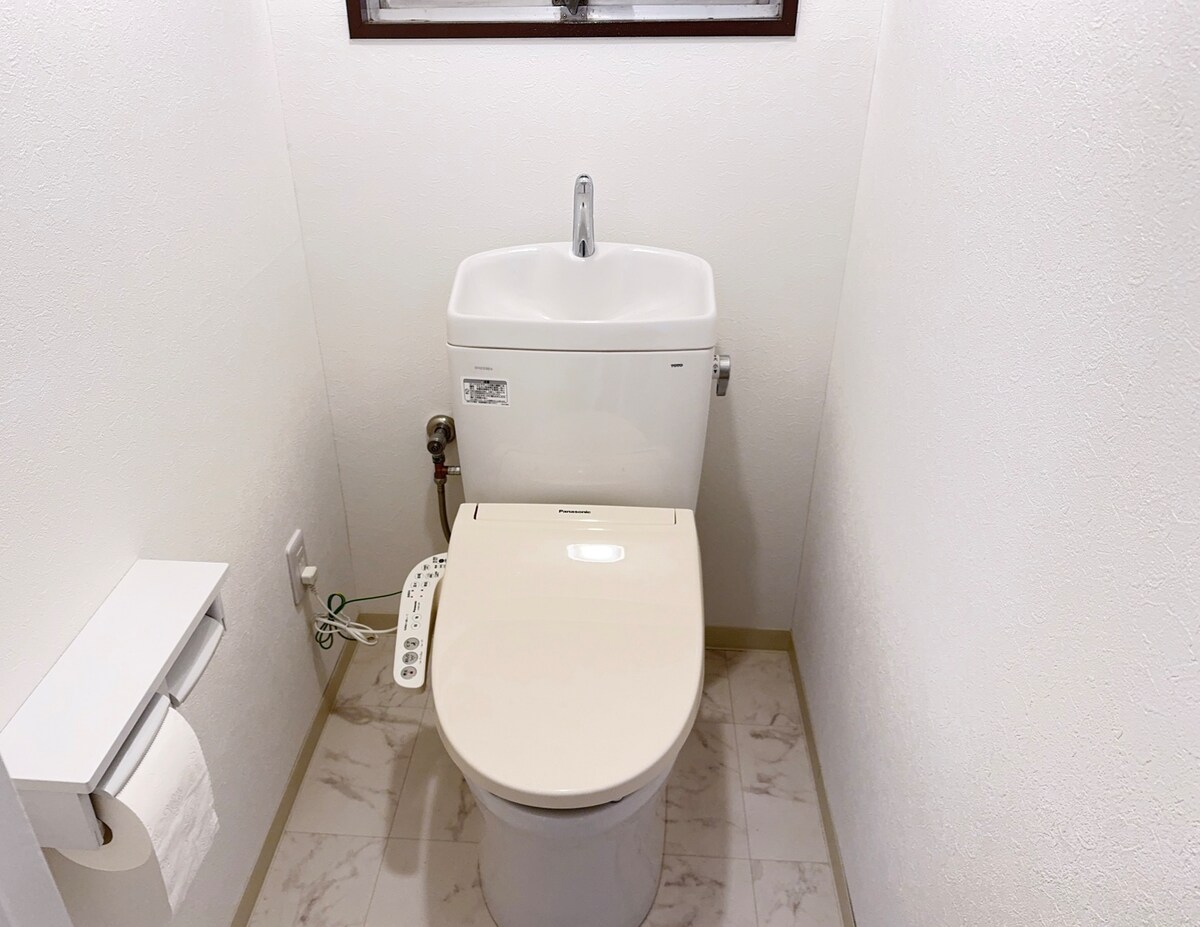 A modern toilet is situated against a clean, white wall in a minimalist bathroom design. The toilet features a sleek design, and a roll of toilet paper is placed on a nearby holder. Neutral tiles cover the floor, enhancing the simplicity of the space.