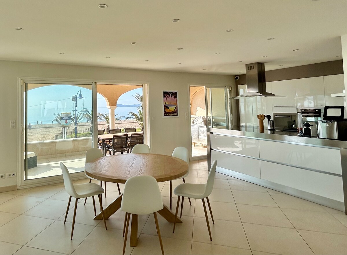 A spacious dining area features a round wooden table surrounded by six white chairs. Large glass doors provide a view of the beach and outdoor seating area, allowing natural light to fill the space. The modern kitchen is equipped with sleek white cabinetry and appliances.