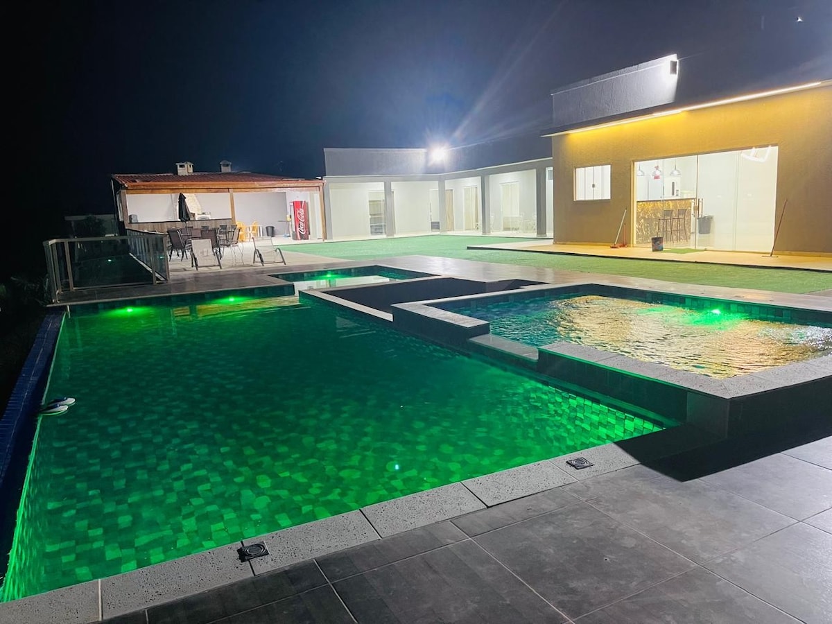 The image showcases a modern outdoor space at night, highlighted by an infinity pool featuring illuminated green lighting. A separate hot tub is visible adjacent to the pool. The background reveals a spacious building, with warm light emanating from large windows.