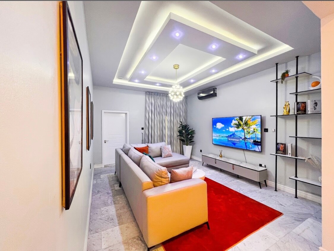 A modern living area features a light-colored sectional sofa with decorative pillows facing a large wall-mounted television. A vibrant red area rug contrasts against the marble flooring. Soft lighting is provided by a stylish ceiling fixture, and sheer curtains frame a window, creating an inviting atmosphere.
