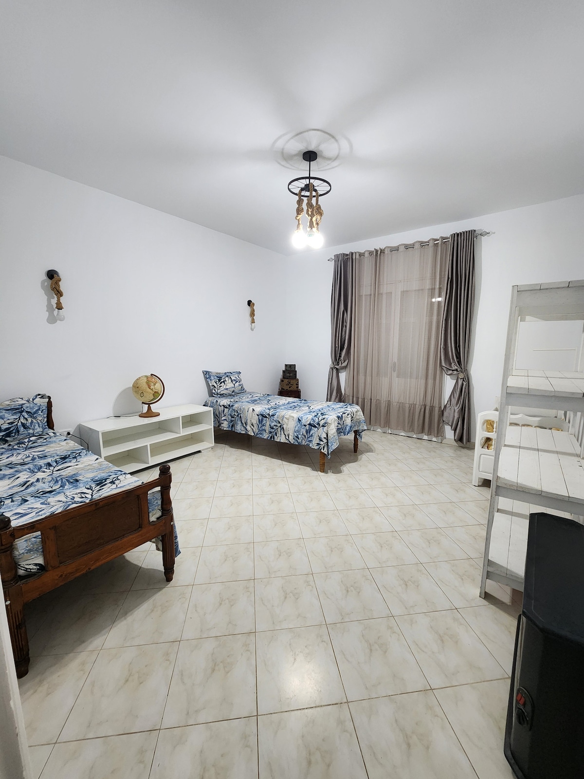 A well-lit room features two twin beds dressed in blue and white floral bedding. A wooden globe sits on a white shelf between the beds. Natural light enters through a large window covered with sheer curtains, and a small white bookcase is positioned against one wall.
