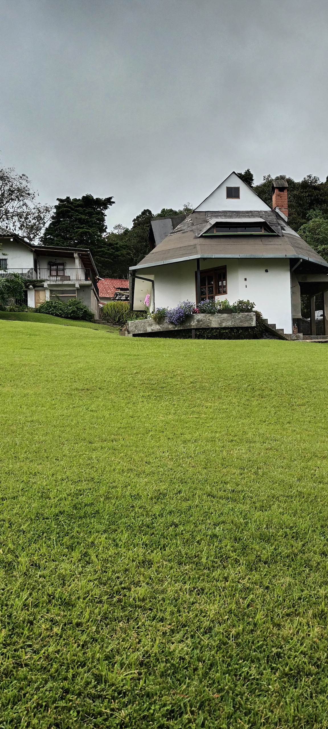 A spacious, grassy lawn surrounds a two-story house with a white exterior and a sloped roof. The home is set against a backdrop of trees, complemented by colorful flower beds near the entrance. A neighboring building adds to the serene environment.