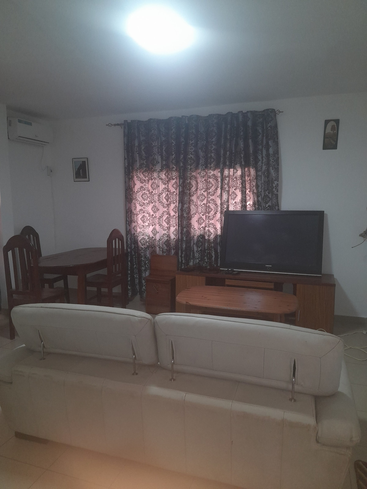 A comfortable living area features a light-colored sofa facing a wooden television unit. A dining table with four dark wooden chairs is positioned nearby, and a window with patterned curtains allows natural light to filter in. A ceiling light provides additional illumination.