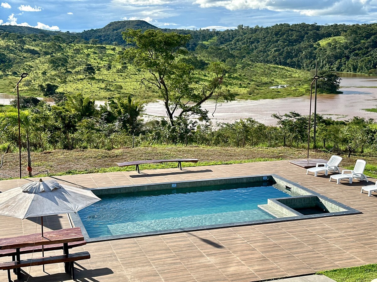 A serene outdoor area features a swimming pool with a connected hydromassage section. Sun loungers are arranged around the pool, while a picnic-style table sits nearby. Lush greenery and rolling hills provide a scenic backdrop, with a river visible in the distance.