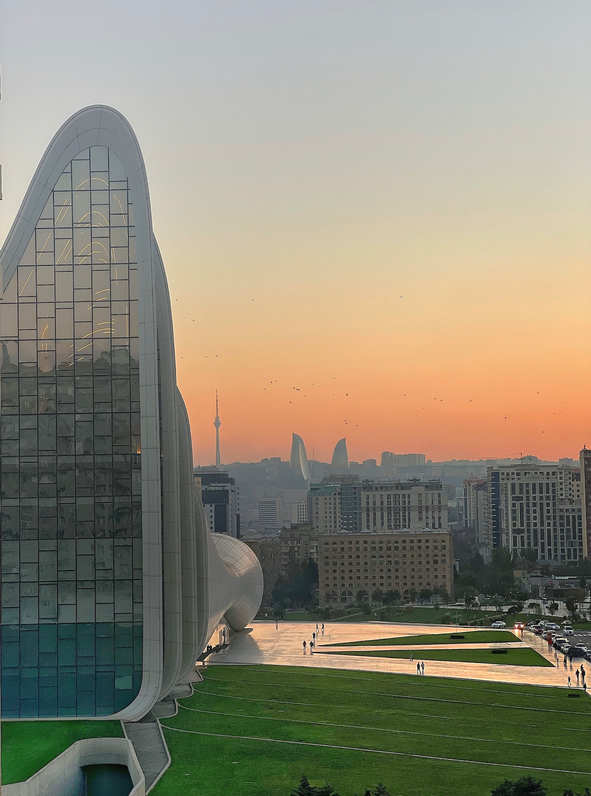 The image features the Heydar Aliyev Center's modern architecture, characterized by sweeping curves and a reflective facade. A sunset casts warm colors over the scene, with silhouettes of distant hills and city buildings visible in the background, including the Flame Towers.
