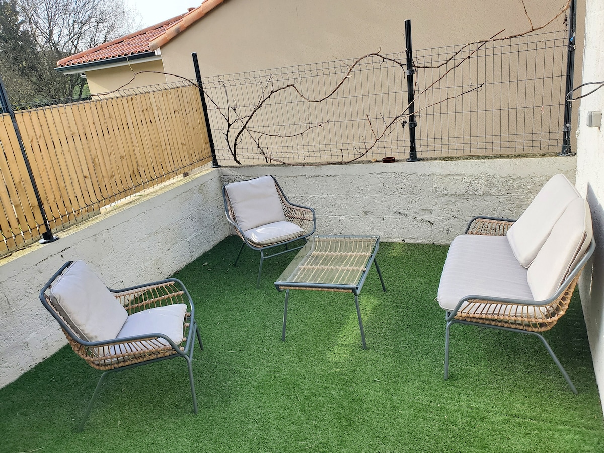 An outdoor seating area features four soft cushioned chairs arranged around a low coffee table. The space is surrounded by a short wall, with a green artificial grass covering providing a comfortable place to relax.