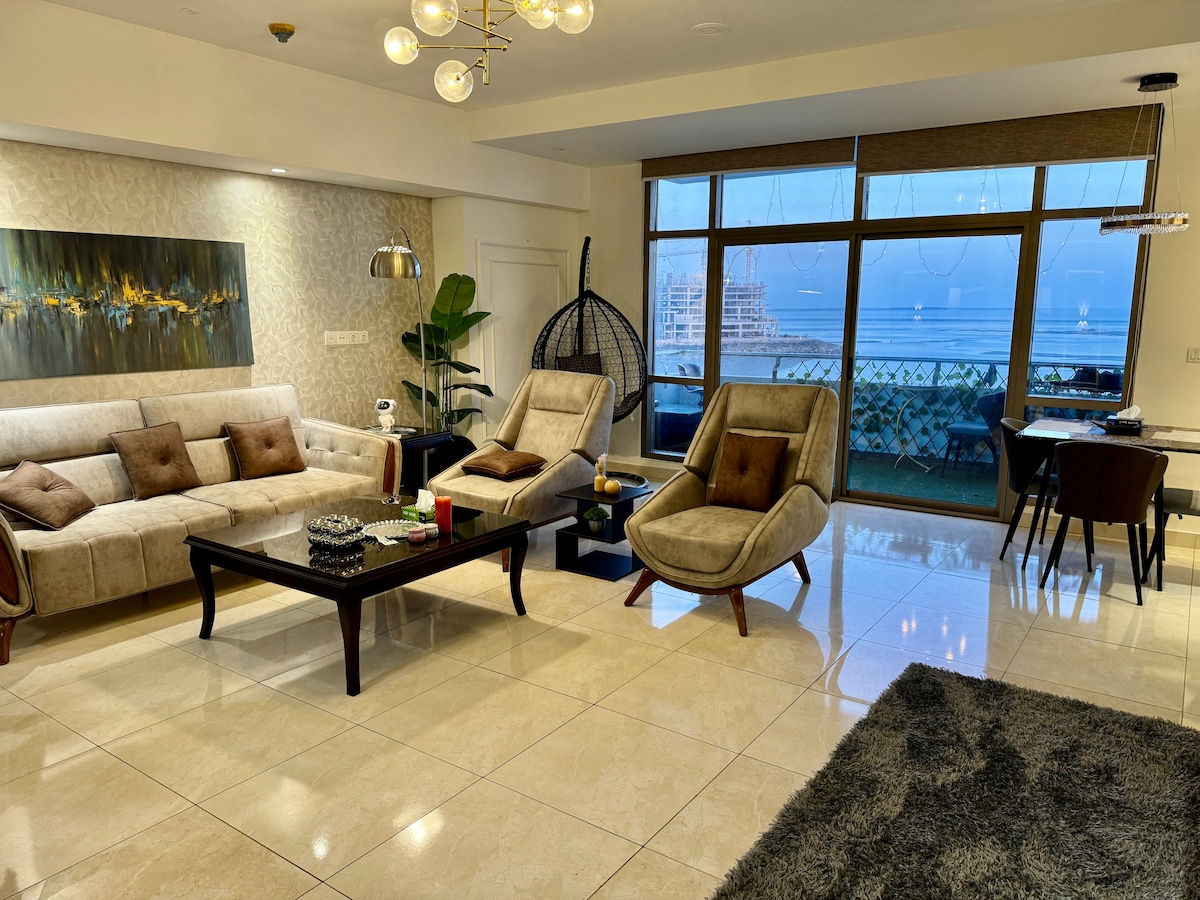 A spacious living area features a modern sectional sofa adorned with decorative cushions. Two stylish lounge chairs complement the decor. Natural light fills the space through large windows, offering views of the sea. A dining area equipped with a sleek table and chairs is visible in the background.