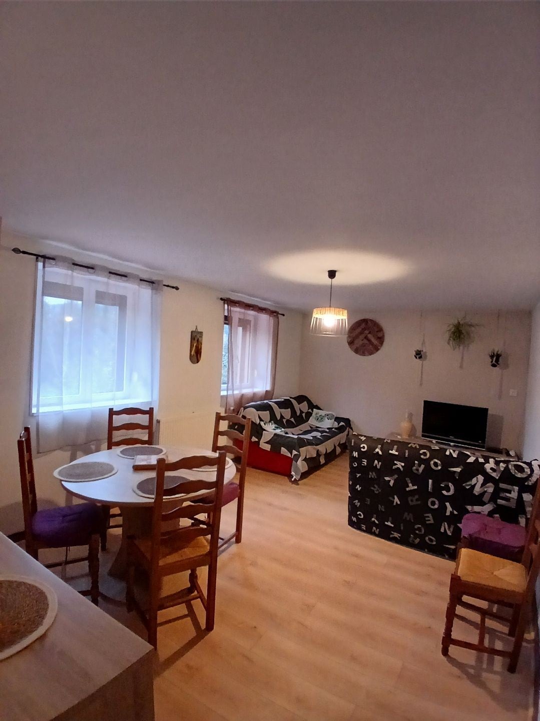 A spacious living area features a dining table set for four, accompanied by wooden chairs. A cozy sofa with dark patterns is positioned next to a television. Large windows allow natural light to brighten the room, enhancing the overall comfort of the space.