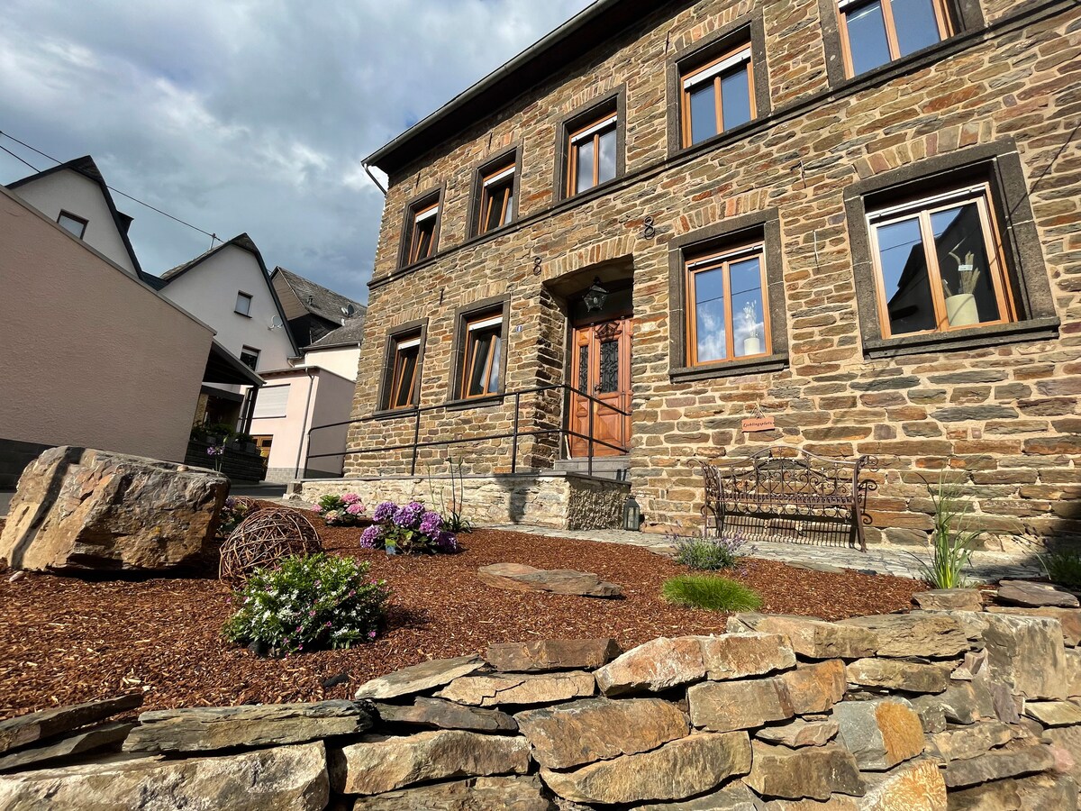 The historic stone house, built in 1887, features multiple windows that allow natural light to illuminate the exterior. A landscaped garden with colorful flowers and decorative elements is visible in the foreground. A stone pathway leads up to the entrance, enhancing the welcoming appeal.