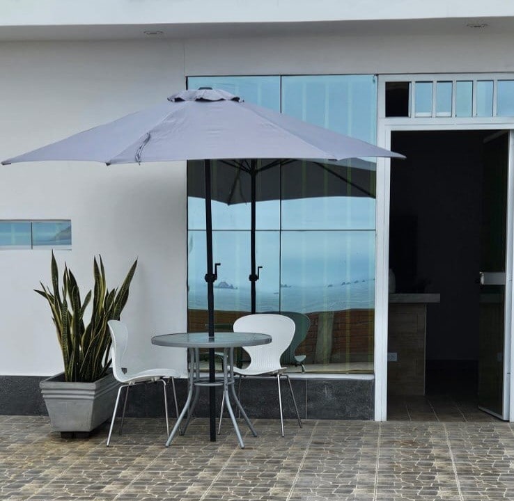 A shaded outdoor seating area features a small round table and two chairs under a large umbrella. A potted plant adds a touch of greenery, while large windows reflect the ocean view, enhancing the connection to the natural surroundings.