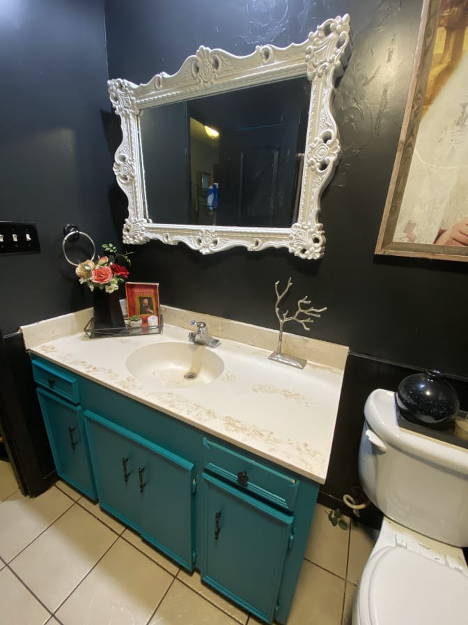 A stylish bathroom features a turquoise vanity with a marble countertop and decorative black hardware. A large ornate mirror hangs above the sink. A small, decorative tree sculpture and a floral arrangement are positioned on the counter, adding artistic elements to the space.