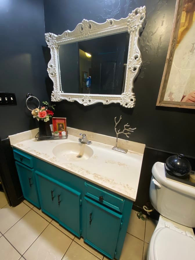A bathroom features a turquoise vanity with a marble countertop and a sink. A large ornate mirror is mounted above the sink, reflecting the space. Decorative elements include a small vase of flowers and a framed picture on the counter. Tiles cover the floor.