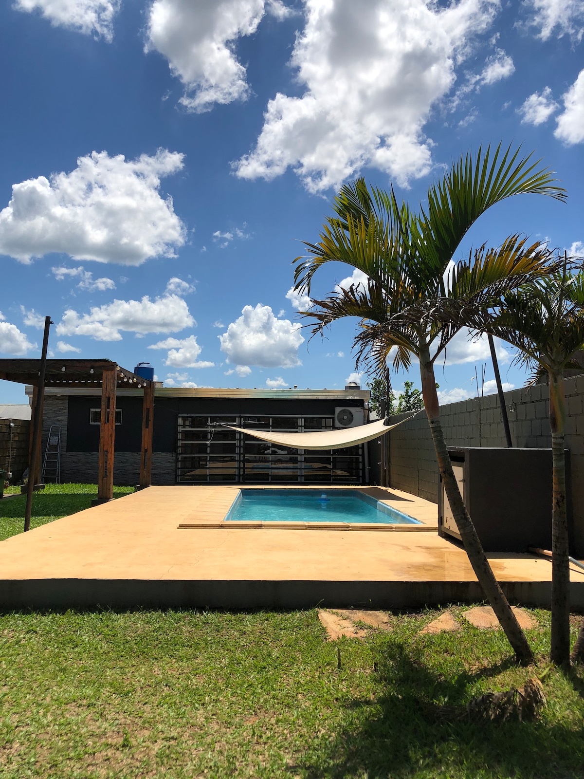 A clear blue sky is overhead, while a small swimming pool is placed on a sunlit patio surrounded by green grass. A hammock is gently draped between wooden posts, and a palm tree adds a touch of greenery to the outdoor space.