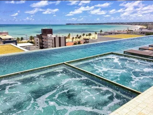 An infinity pool extends towards the horizon, blending with the vibrant blue sea. The view encompasses sandy shores and palm trees along the coastline, with distant buildings adding a scenic backdrop under a clear sky.