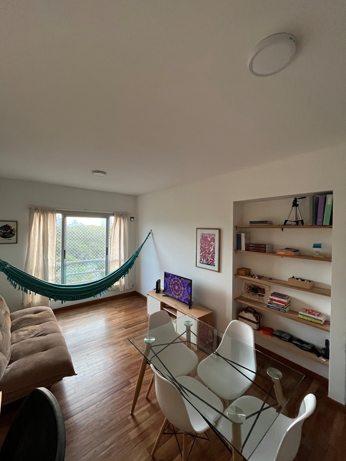 A bright living space features a comfortable sofa, a hammock suspended by the window, and a glass dining table surrounded by white chairs. Natural light streams in through the large window adorned with sheer curtains, illuminating the minimalist decor and a shelving unit with various items.