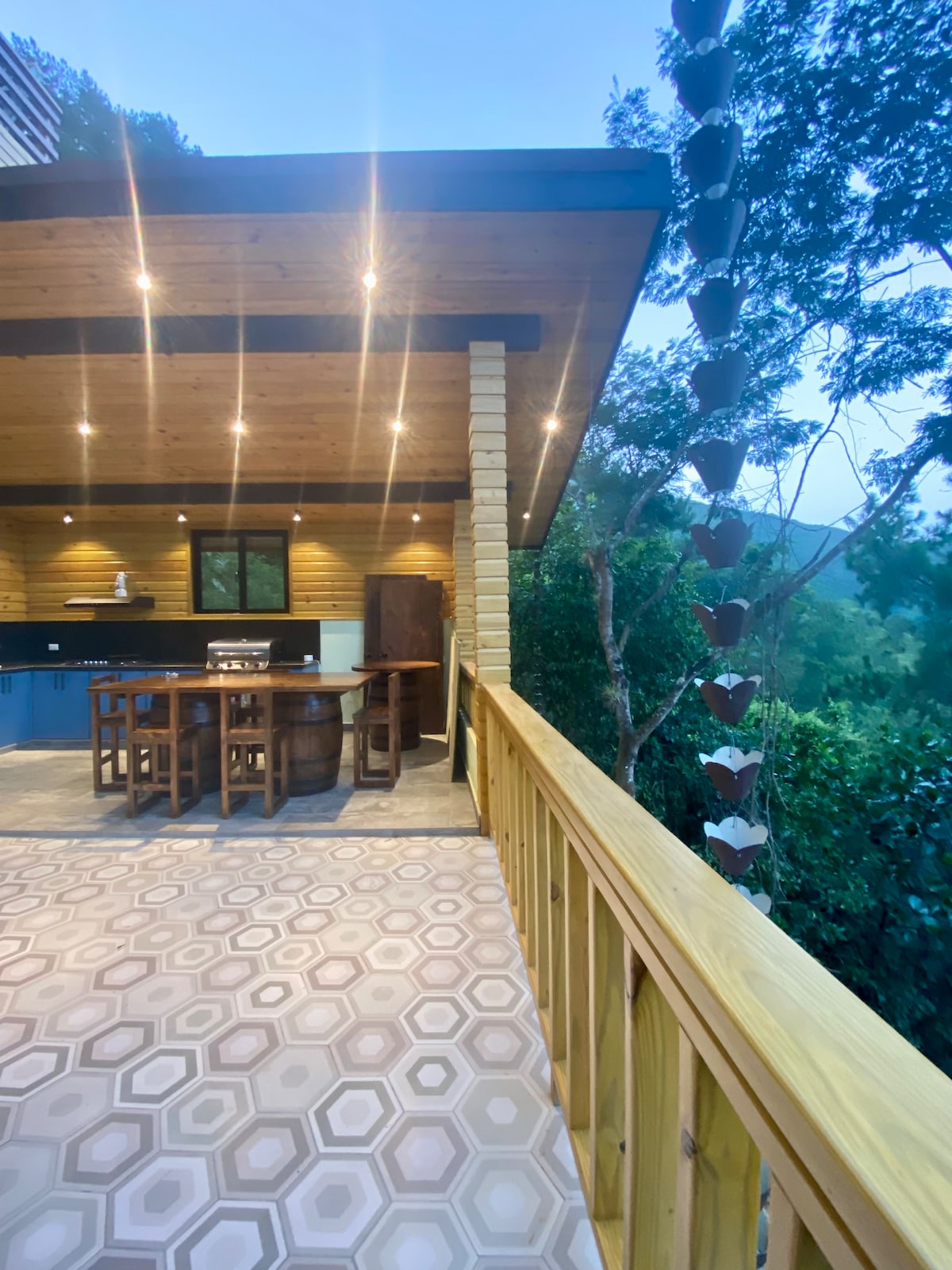 An outdoor dining area features a wooden table surrounded by matching chairs, set under a spacious wooden roof. Soft lighting illuminates the space, with lush greenery visible in the background, enhancing the natural setting. The flooring showcases a geometric tile pattern.