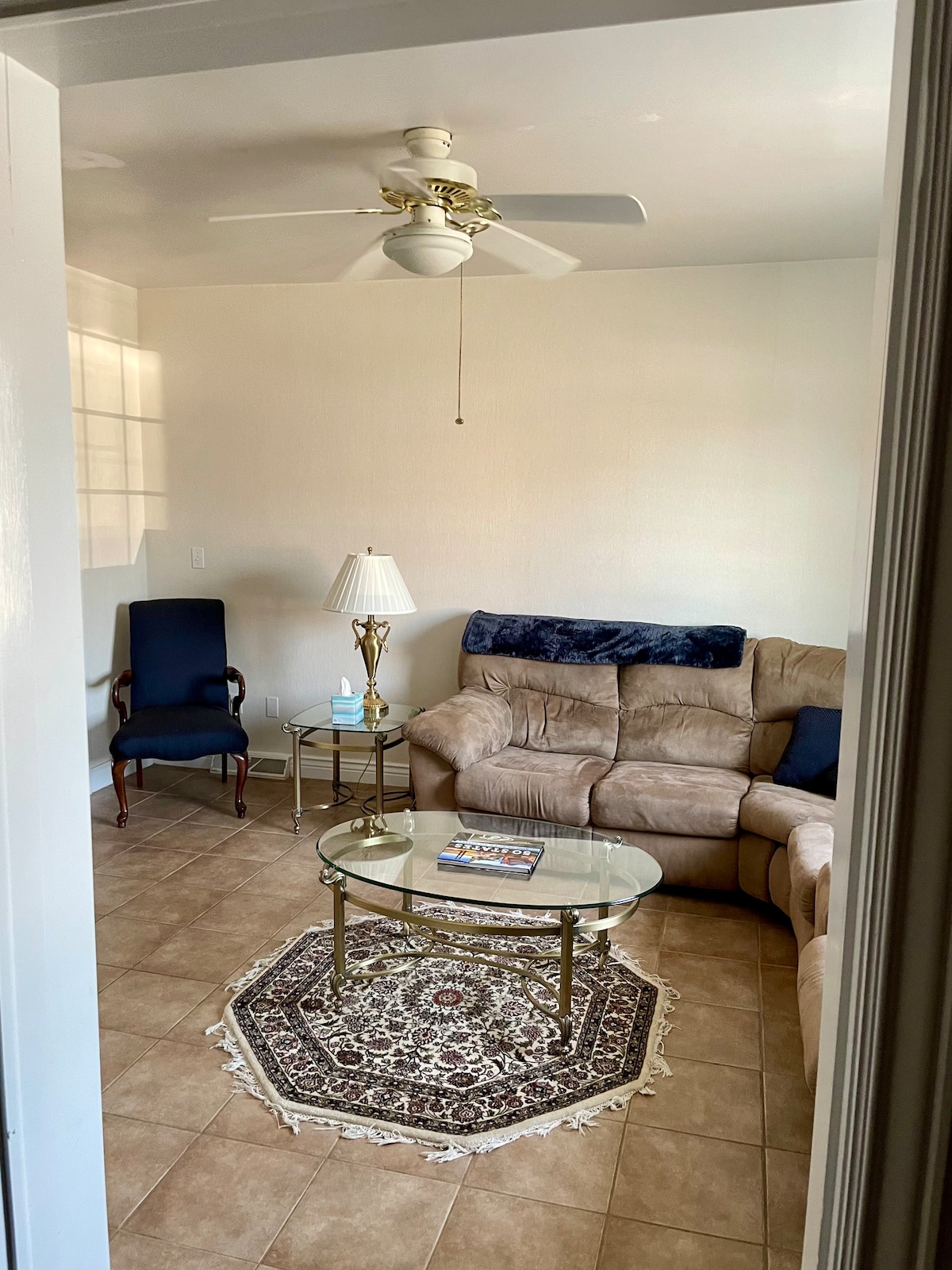 A cozy living area is displayed with a sectional sofa in tan upholstery. A glass coffee table sits on a patterned area rug. A table lamp is positioned beside the sofa, while a single armchair is placed in the corner. Neutral tones enhance the relaxed atmosphere.