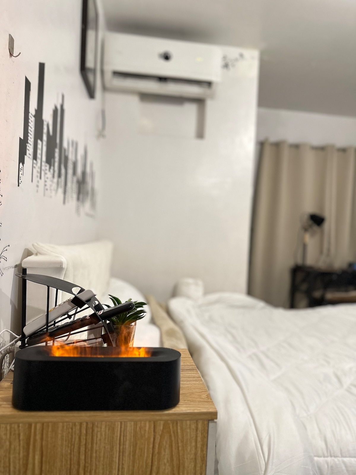 A neatly made bed is positioned against a light-colored wall adorned with a city skyline design. A bedside table holds a small plant and a modern lamp. An air conditioning unit is mounted above, and light curtains softly frame a window.