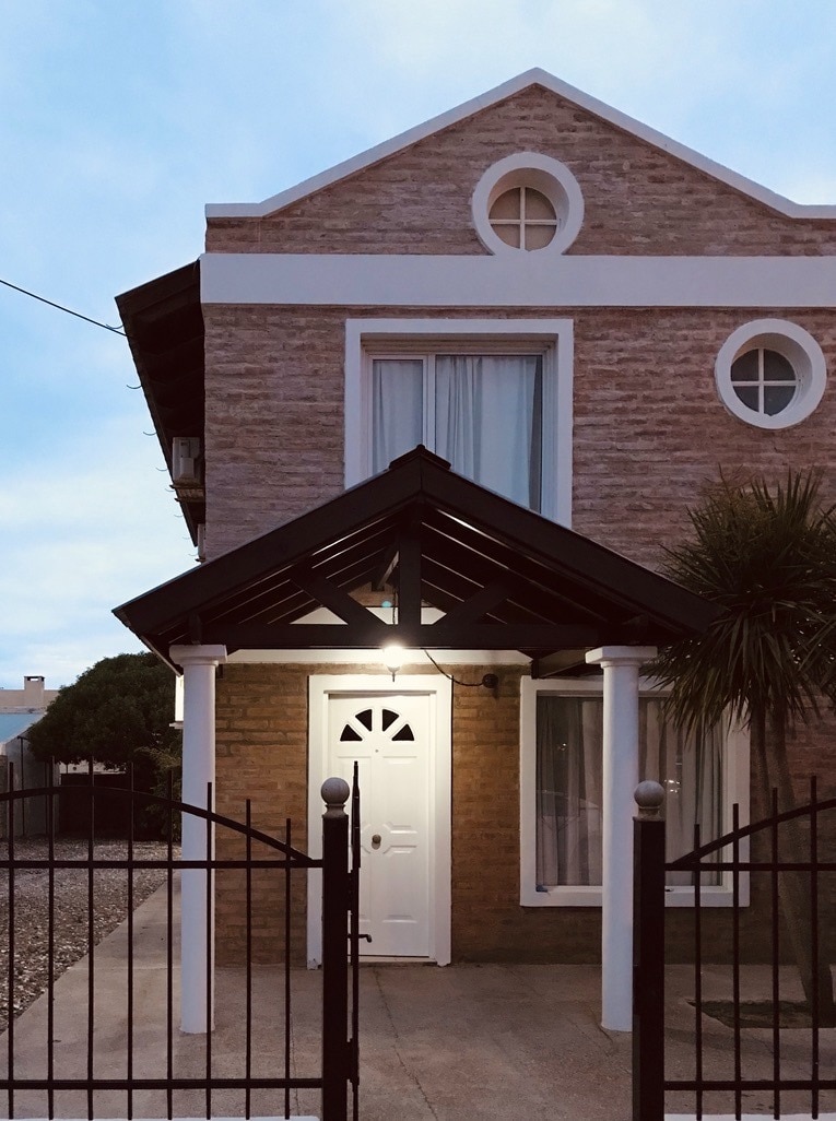 The entrance to the house features a gabled roof supported by white columns. A well-lit door with a circular window invites guests inside. The warm-colored bricks contrast with the soft blue sky, while a garden area is visible to the side.