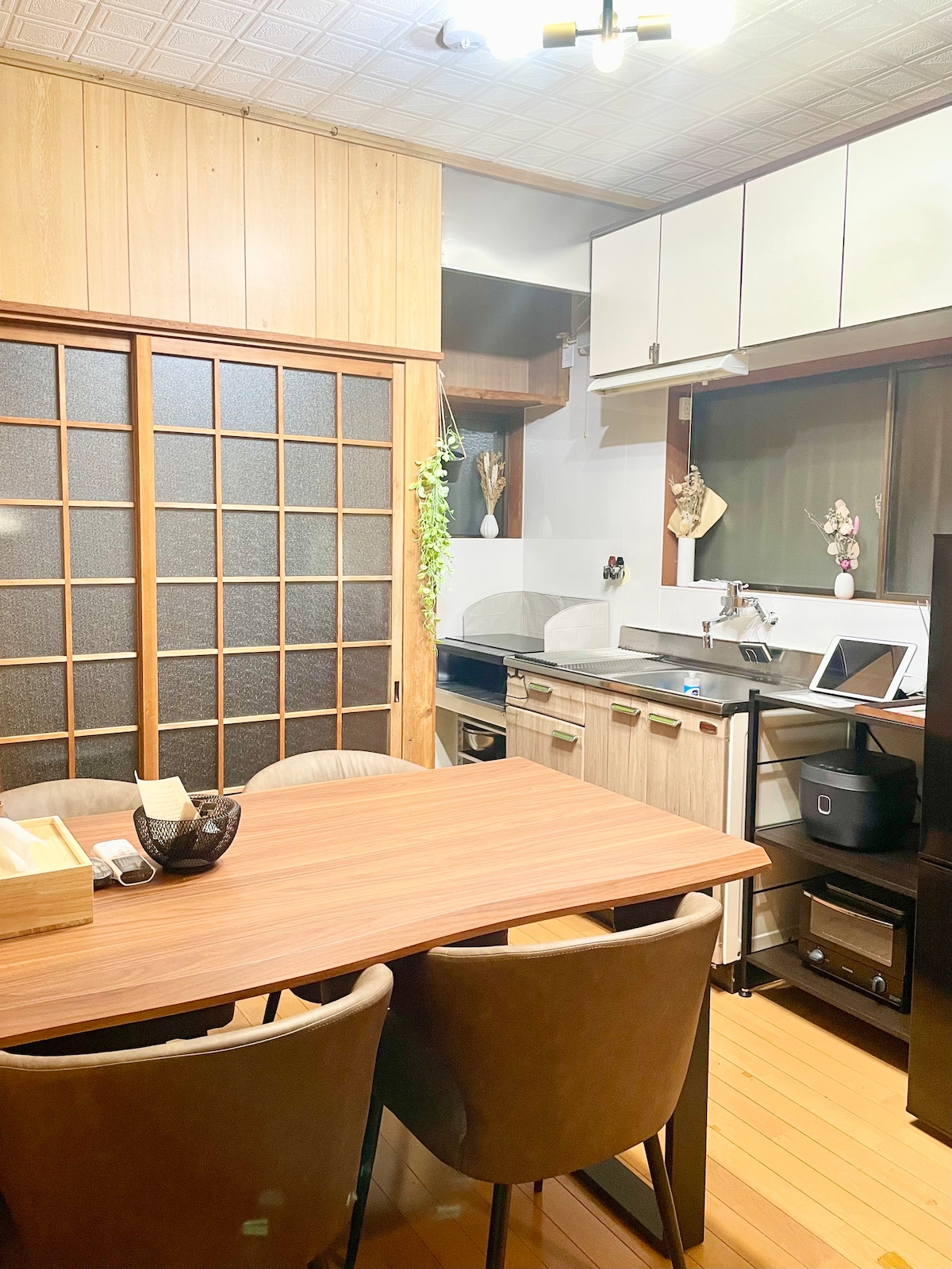 The dining kitchen area is depicted, featuring a wooden table with four chairs. Cooking appliances, including an IH stove, are arranged in a modern kitchen. Light wood cabinetry reflects simplicity, while sliding glass doors provide access to natural light.