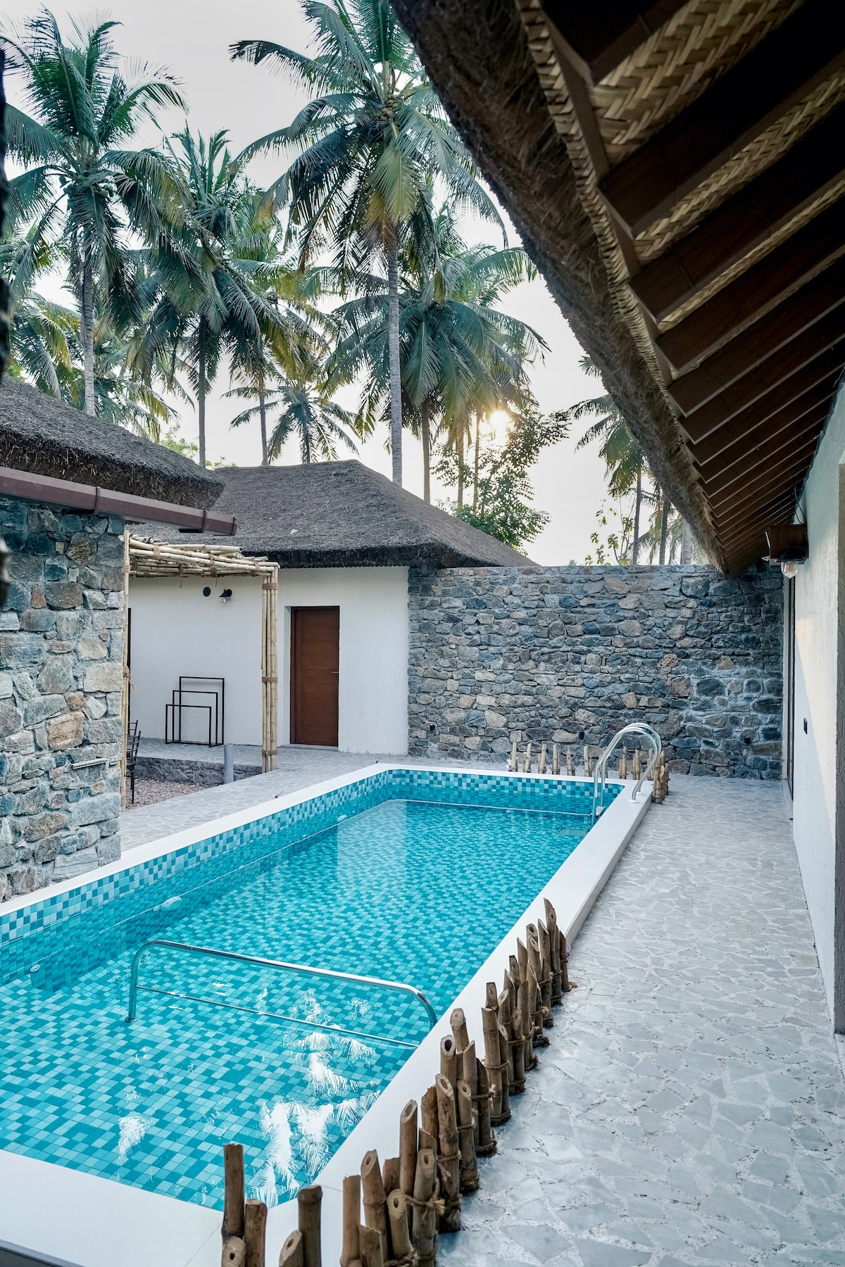 A private pool is surrounded by stone walls and thatched-roof structures, offering a secluded ambiance. Tall palm trees rise in the background, while the pool features a blue mosaic tile design. A stone pathway leads to the pool area, enhancing the retreat's tranquil setting.