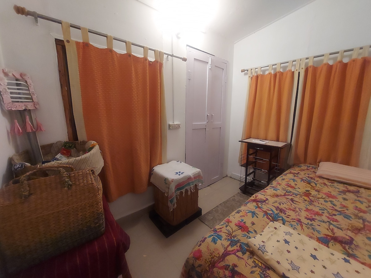 The room features a spacious bed with a colorful patterned quilt. Soft orange curtains frame the windows, allowing natural light to filter in. A small side table is placed beside the bed, while woven baskets and a chest provide additional storage and texture.