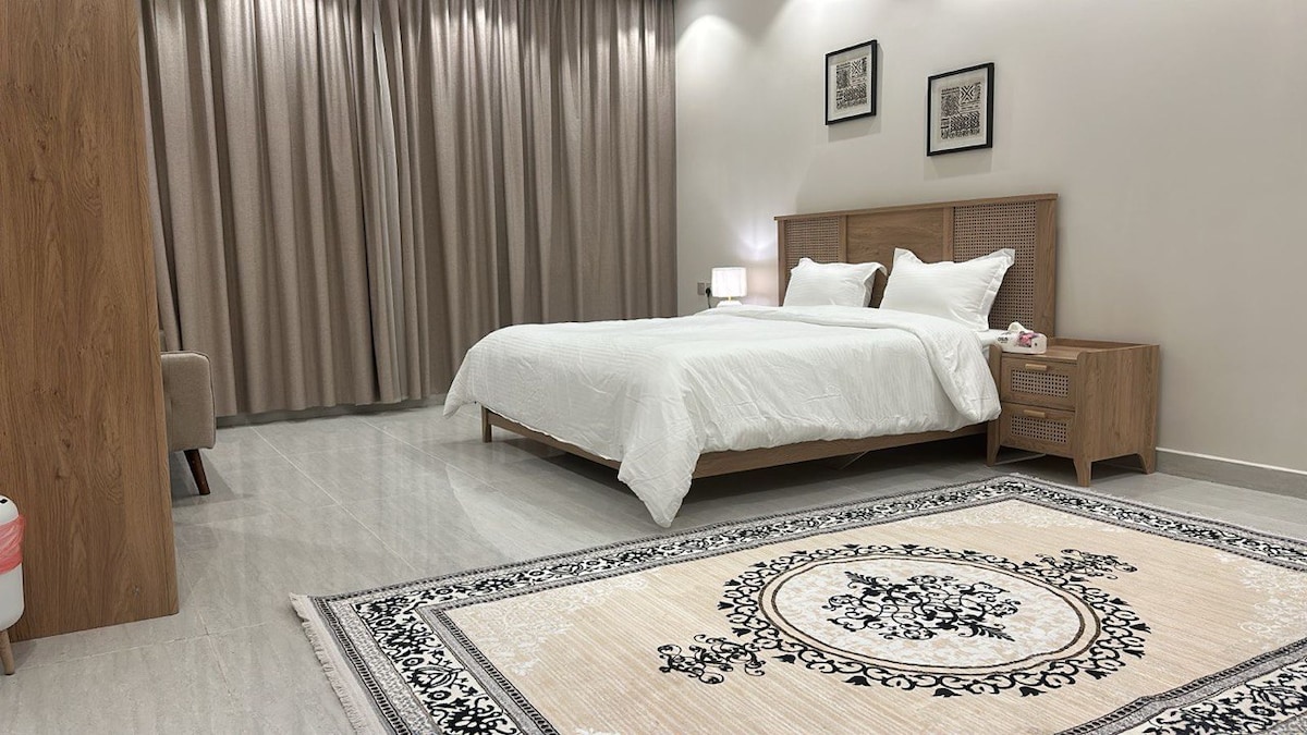 A spacious bedroom features a large, light-colored bed with crisp white linens. A decorative area rug adds texture to the floor. Soft, neutral-toned curtains frame the large window, and two framed artworks hang above the bed, contributing to the modern aesthetic.