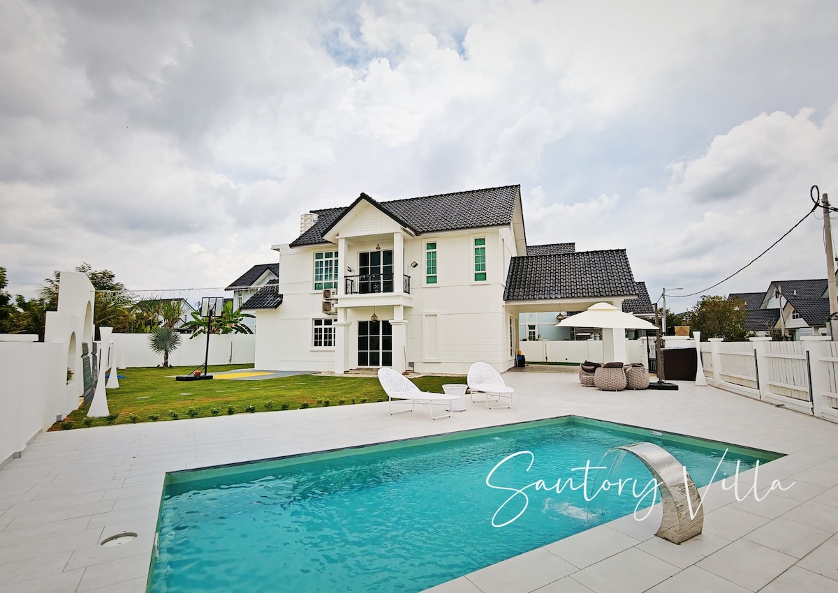 The exterior of Santory Villa is presented, featuring a private swimming pool with clear water. Sun loungers are positioned nearby on the tiled patio. Lush green grass surrounds the house, which displays a blend of modern architecture with large windows and a balcony on the upper floor.