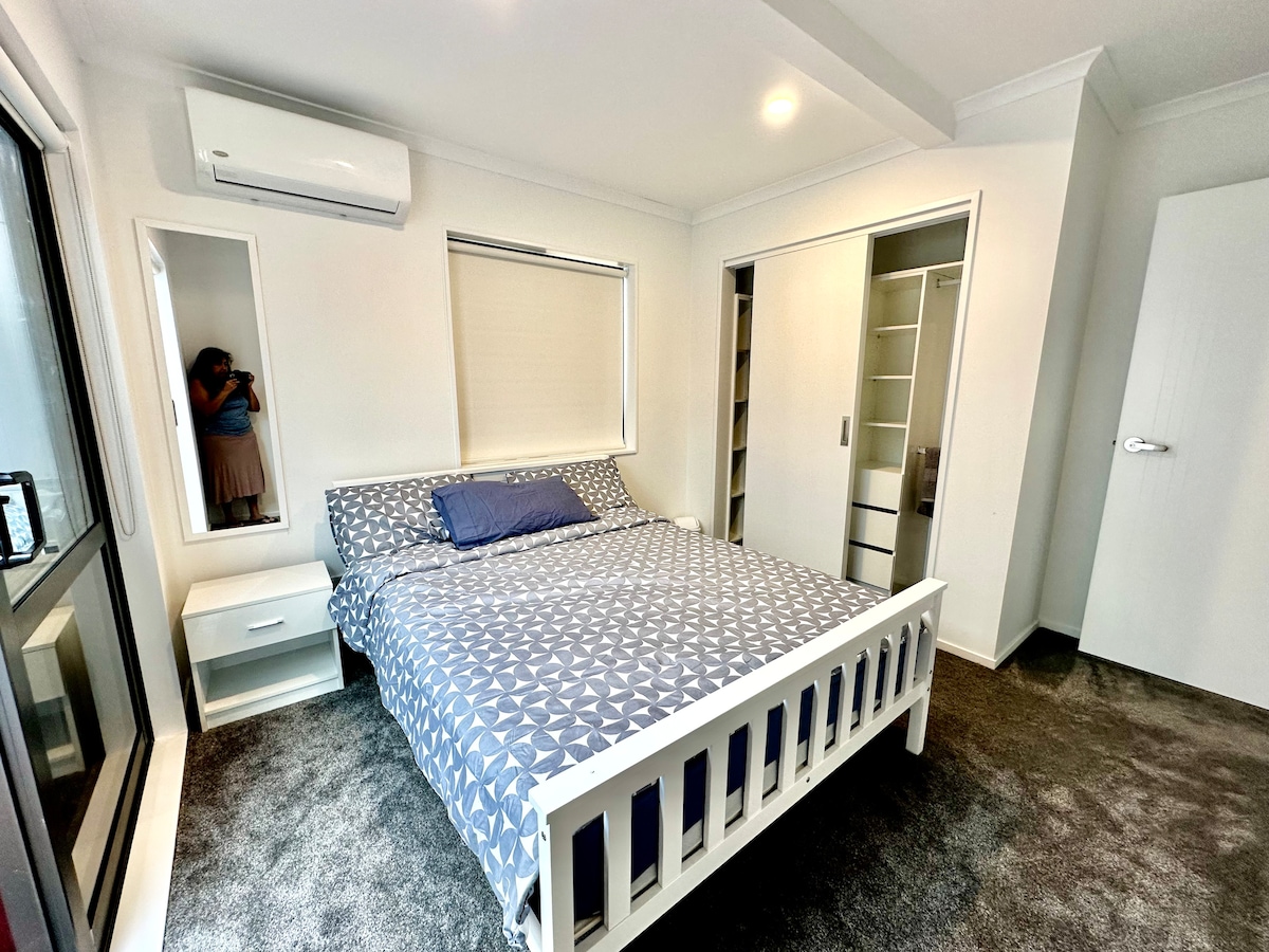 A well-lit sleeping area features a spacious bed with a geometric patterned blanket and a coordinating pillow. Soft, plush carpet covers the floor. A large window allows natural light to enter, while a built-in closet is positioned on one side. An air conditioning unit is visible on the wall.