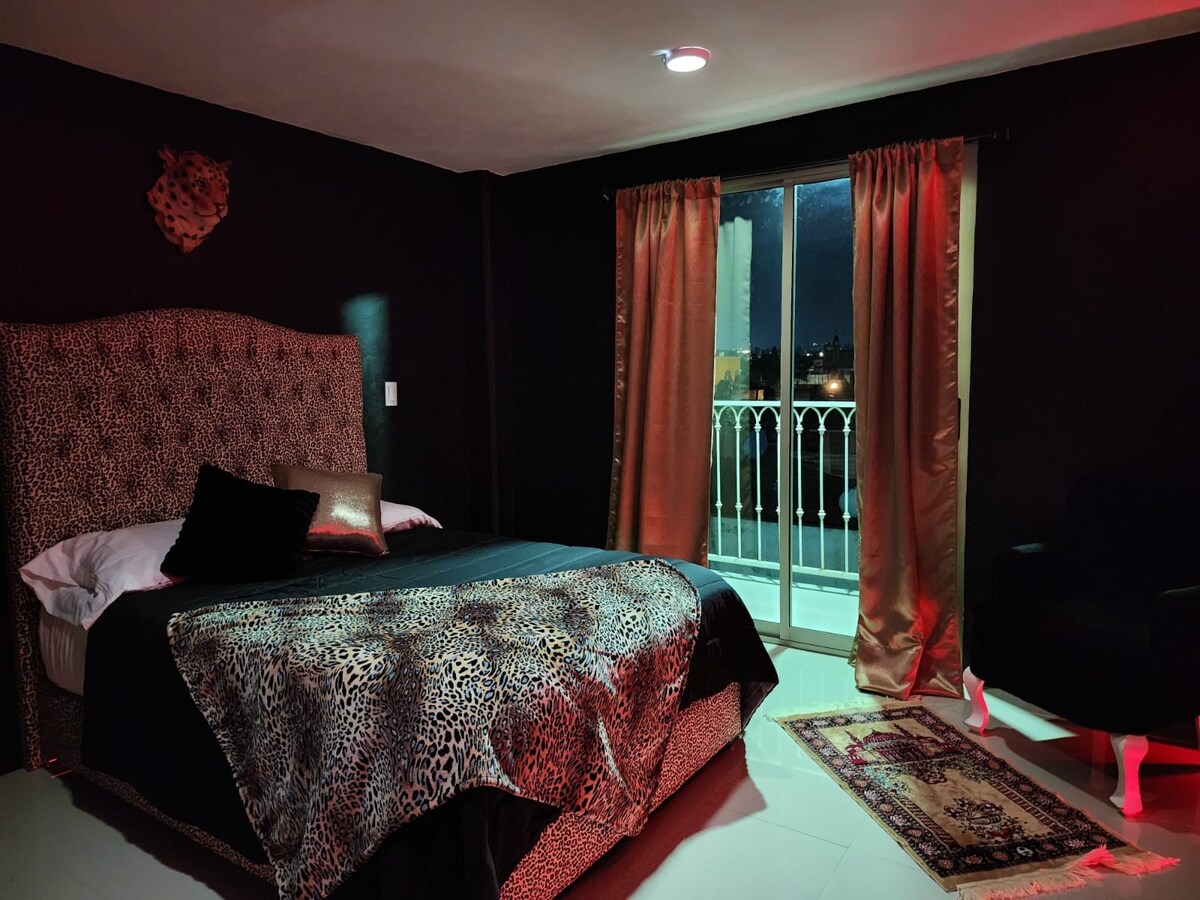 A stylish room features a spacious bed with a leopard print headboard and bedding. Soft lighting highlights the bold, dark walls. Sheer curtains frame the balcony doors, which offer views outside. A plush chair and a decorative rug complete the inviting and elegant decor.