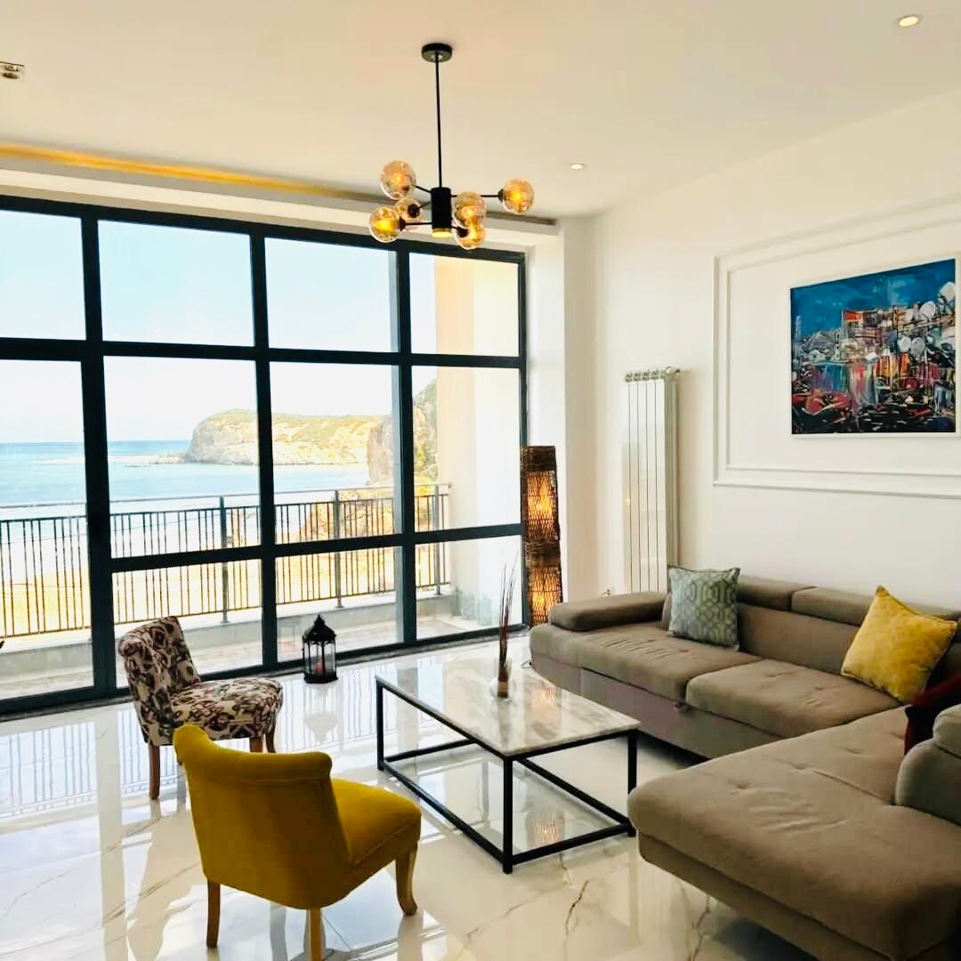 A bright living area features a large window with an ocean view, allowing natural light to fill the space. A sofa is complemented by two accent chairs, while a glass coffee table rests on a glossy floor. Art decorates the walls, adding visual interest.
