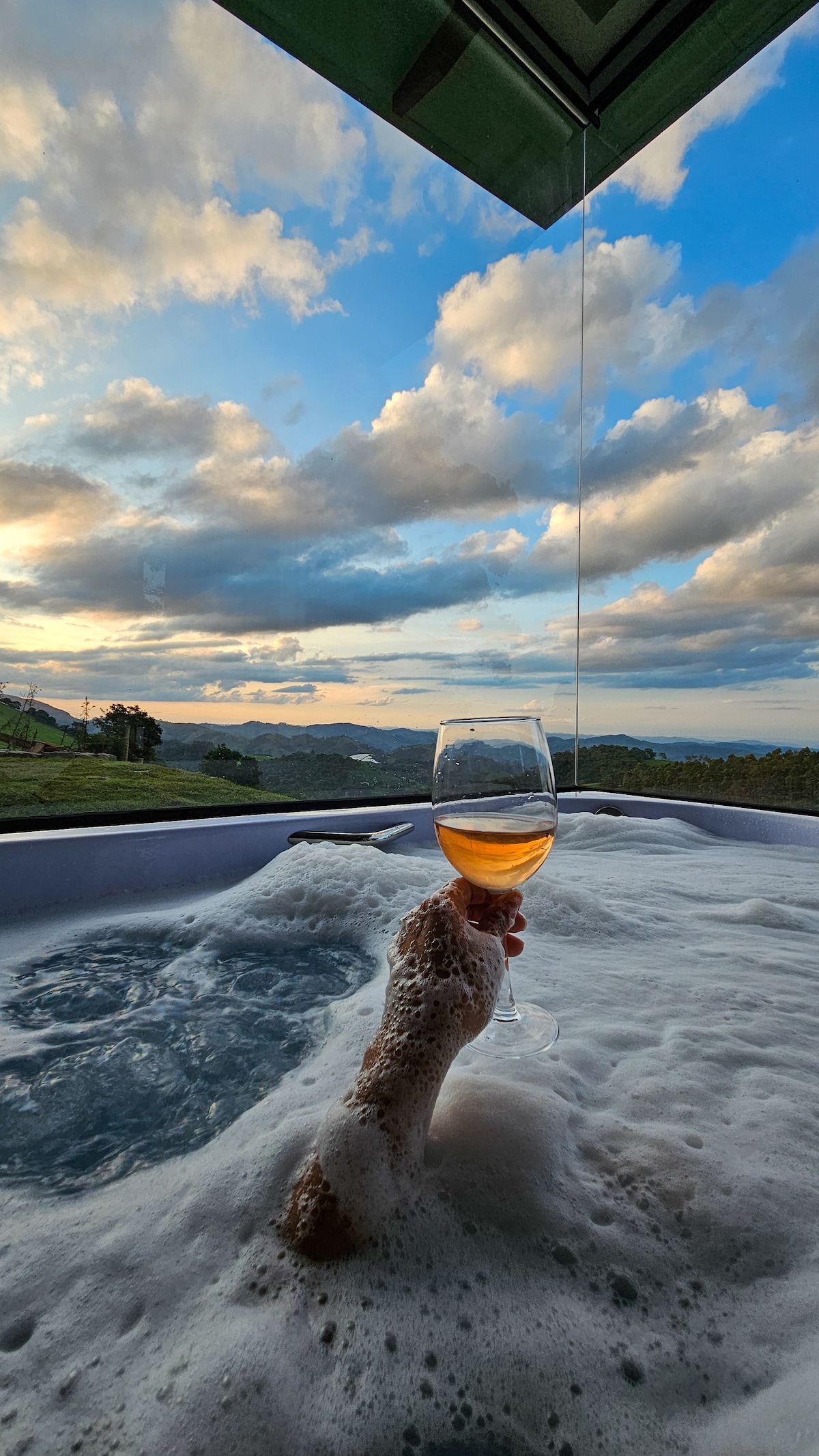 A hand holding a glass of beverage is emerging from a bubbly hot tub, with a scenic view of rolling hills under a partly cloudy sky. Soft hues of sunset light fill the background, creating a serene atmosphere.