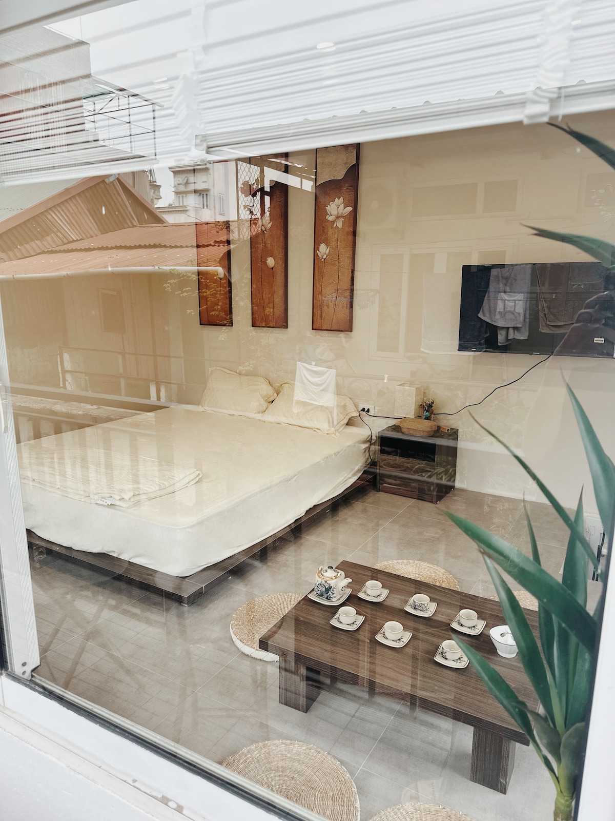 A room is viewed through a large window, revealing a spacious bed and a low wooden table set with cups. Decorative wall art complements the light-colored walls, while a television is mounted across from the bed. Natural light fills the area, enhancing the inviting atmosphere.