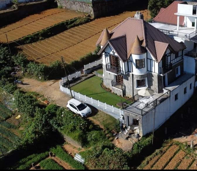 An aerial view of a charming villa set amidst cultivated fields. The building features a distinct peaked roof and is surrounded by lush greenery. A well-maintained lawn and outdoor seating area are visible, along with a parked car in the driveway.