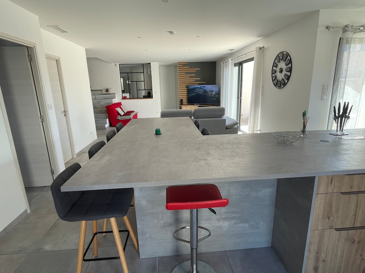 An open-concept kitchen area features a large, gray countertop with bar stools, offering a modern feel. The space is well-lit, showcasing a comfortable seating arrangement with a sofa and a red chair in the background. Large sliding doors provide access to outdoor views.