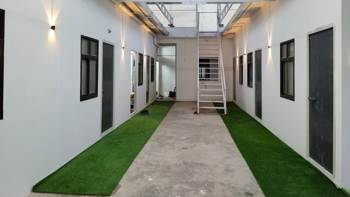 A well-lit corridor features a smooth concrete floor with artificial grass along the sides. Doors lead to individual rooms, and a staircase is visible at the far end, providing access to an upper level. Wall-mounted lights enhance the overall brightness.
